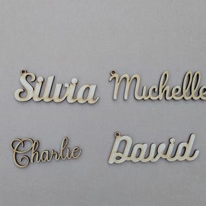 May include: Four wooden name cutouts: "Silvia", "Michelle", "Charlie", and "David". Each name is in a cursive font, with a small hole at the top for hanging. The wood has a natural, light color.