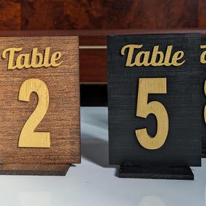 May include: Four table number signs with a wooden and black finish. The signs have the word "Table" and a number on them. The numbers are 2, 5, 8, and 9.