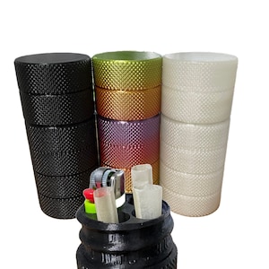 Op de afbeelding: Een zwarte, meerlaags opbergcontainer met een aansteker en rolletjes papier erin. De container heeft een getextureerd oppervlak en is open om de inhoud te laten zien. Drie andere containers zijn zichtbaar op de achtergrond, een groene, een gouden en een witte.