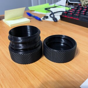 May include: Two black 3D-printed cylindrical objects with textured bases. One is a complete cylinder with a ridged top, and the other is a separate lid. The objects are on a light brown wooden surface.