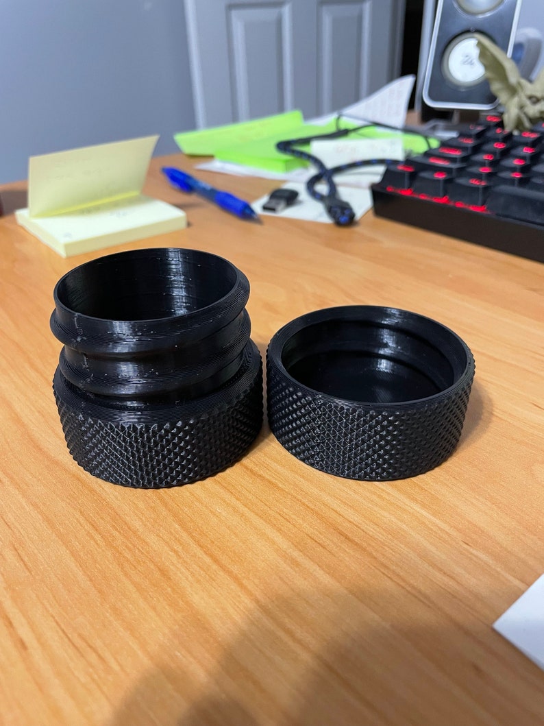 May include: Two black 3D-printed cylindrical objects with textured bases. One is a complete cylinder with a ridged top, and the other is a separate lid. The objects are on a light brown wooden surface.