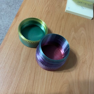 May include: Two small, round containers with textured sides. One is green and gold, the other purple and red. The containers are on a light brown wooden surface. A sticky note with handwritten numbers is in the background.