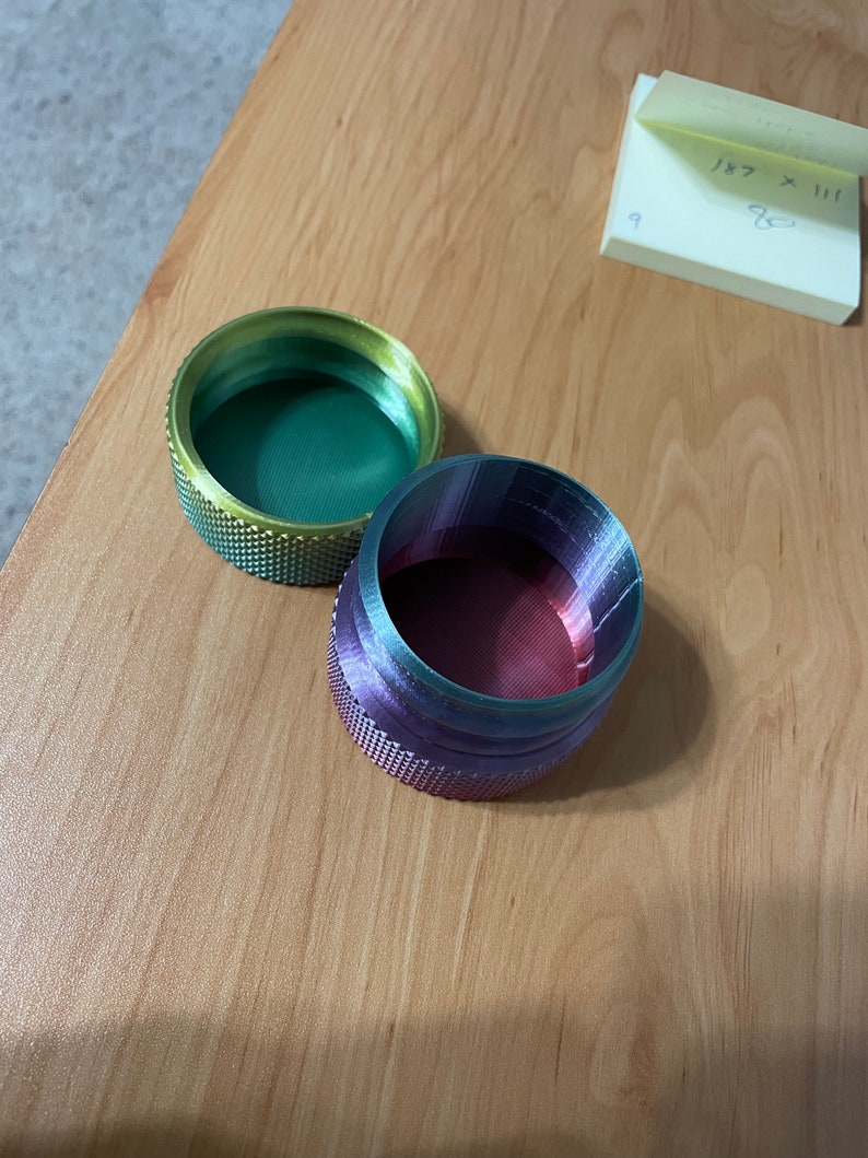 May include: Two small, round containers with textured sides. One is green and gold, the other purple and red. The containers are on a light brown wooden surface. A sticky note with handwritten numbers is in the background.