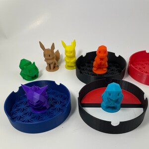 May include: Five colorful 3D printed Pokemon figurines on top of four different colored ashtrays. The ashtrays are black, red, blue, and white with black.
