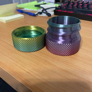 May include: Two 3D-printed objects with textured surfaces. One is a cylindrical shape with a gradient of yellow to green. The other is a taller, multi-tiered shape with a gradient of purple to green. Both objects are on a wooden surface.
