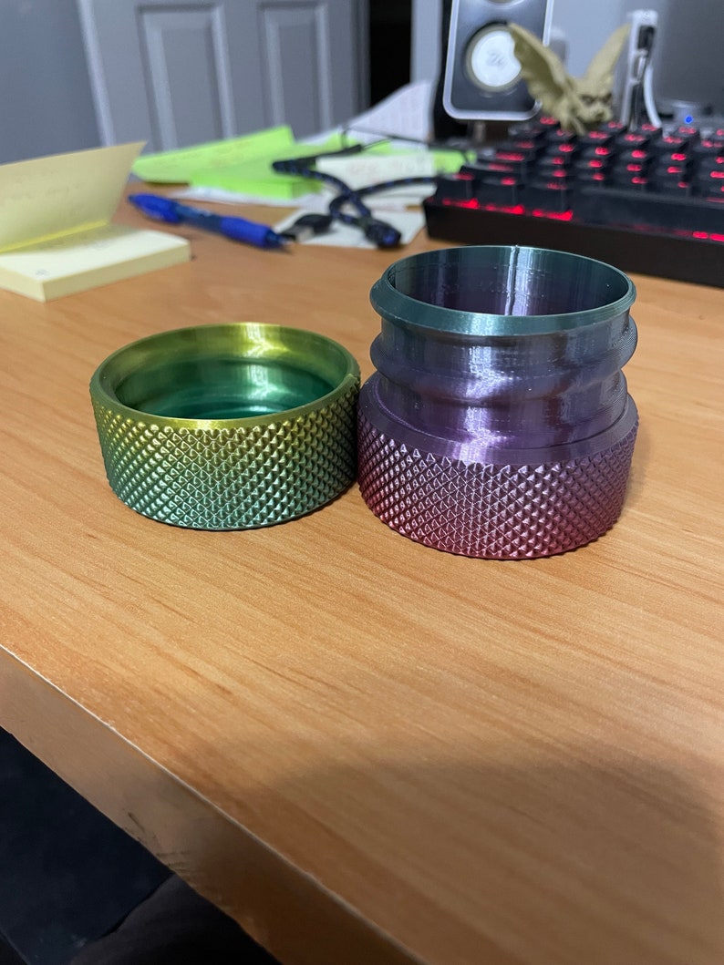 May include: Two 3D-printed objects with textured surfaces. One is a cylindrical shape with a gradient of yellow to green. The other is a taller, multi-tiered shape with a gradient of purple to green. Both objects are on a wooden surface.