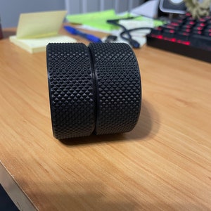 May include: Two black, cylindrical objects with a textured, diamond-patterned surface. The objects are side-by-side, resting on a light brown wooden surface. The objects appear to be components of a larger item.