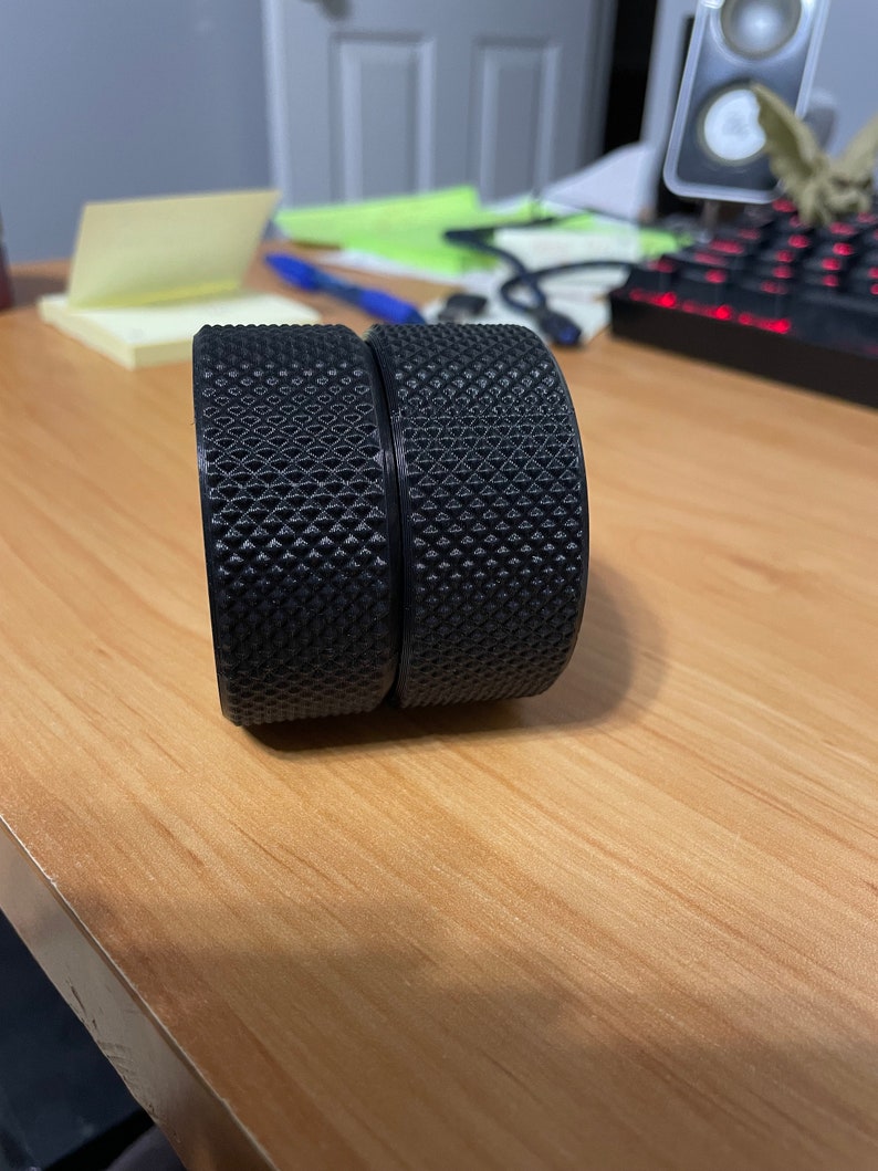 May include: Two black, cylindrical objects with a textured, diamond-patterned surface. The objects are side-by-side, resting on a light brown wooden surface. The objects appear to be components of a larger item.