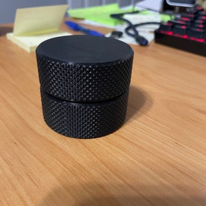 May include: A black, cylindrical object with a textured, diamond-patterned surface. The object is composed of two stacked, identical pieces. The item is sitting on a light brown wooden surface.