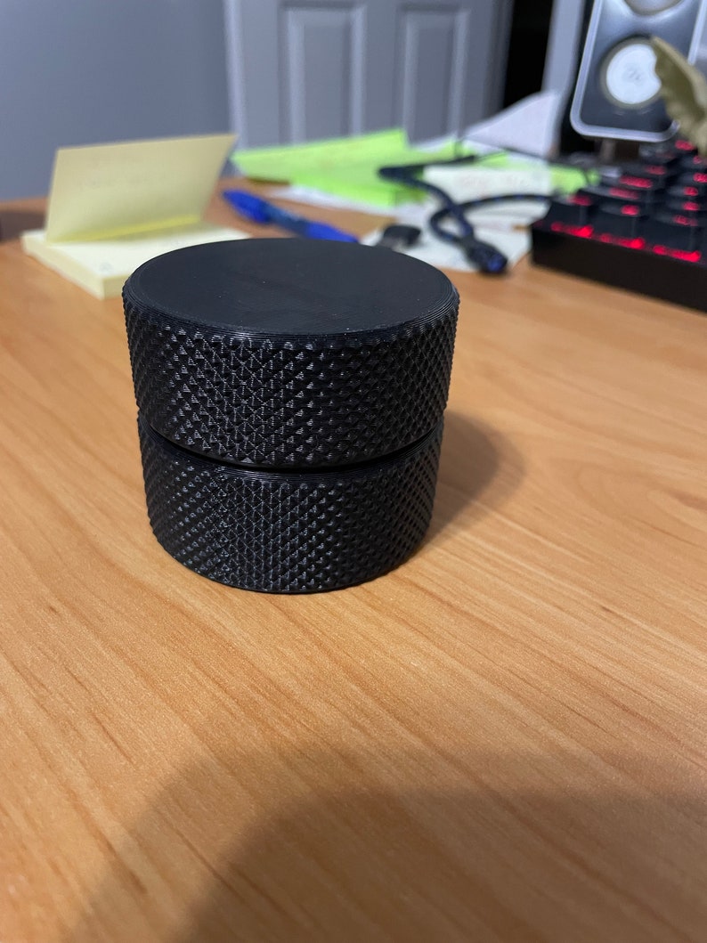 May include: A black, cylindrical object with a textured, diamond-patterned surface. The object is composed of two stacked, identical pieces. The item is sitting on a light brown wooden surface.