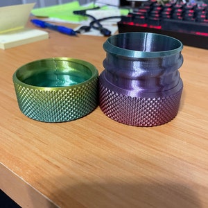 May include: Two gradient-colored, textured cylindrical objects sit on a wooden surface. One is a low, wide cylinder with green and yellow hues. The other is taller, with a purple and green gradient. Both have a diamond pattern.