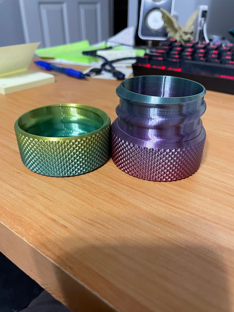 May include: Two gradient-colored, textured cylindrical objects sit on a wooden surface. One is a low, wide cylinder with green and yellow hues. The other is taller, with a purple and green gradient. Both have a diamond pattern.