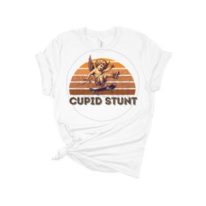 Puede incluir: Camiseta blanca con un gráfico retro de un querubín patinando con el texto "Cupid Stunt" debajo.