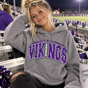 Könnte beinhalten: Ein graumelierter Kapuzenpullover mit einem violett-weißen Grafikdruck, der VIKINGS zeigt, wobei eine Football-Illustration den Buchstaben I ersetzt. Das Design umfasst fünf kleine Sterne über dem Text und eine Kängurutasche.