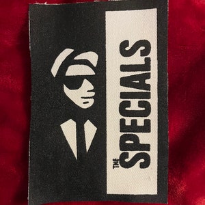 May include: A black and white patch featuring a stylized image of a man in a hat and sunglasses. The text "THE SPECIALS" is printed vertically on the right side of the patch. The patch is rectangular and has a fabric texture.