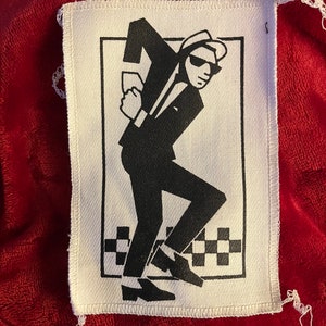 May include: White fabric banner with a black and white illustration of a man in a suit, hat, and sunglasses, in a dancing pose. The banner has a checkered pattern at the bottom and white stitching around the edges. The banner is on a red background.