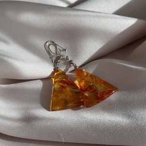 May include: A pair of amber earrings with a triangular shape and silver hooks. The earrings are on a white fabric background.