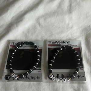 Puede incluir: Dos pulseras de cuentas blancas y negras con las letras "XO" y "OTWOD" en cuentas blancas. Las pulseras se muestran en tarjetas con el texto "The Weeknd" y "Echoes Of Silence".