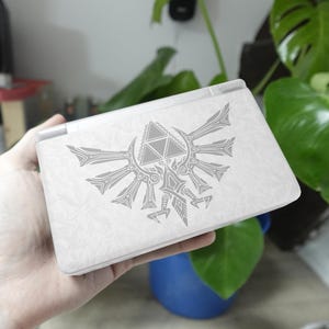Könnte beinhalten: Eine weiße tragbare Spielkonsole mit einem hellgrauen Triforce- und Vogelwappen auf der Oberseite. Die Oberfläche weist ein dezentes, sich wiederholendes florales Muster auf dem gesamten äußeren Gehäuse des rechteckigen Geräts auf.