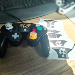 May include: A black video game controller featuring a gold Triforce emblem on the center panel, accompanied by four white adhesive decals displaying various gaming symbols, including a Smash Bros logo and a flaming icon, arranged in a vertical row.