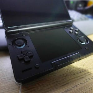 Könnte beinhalten: Eine schwarze Handheld-Spielkonsole mit einem aufklappbaren Dual-Screen-Design. Das Gerät verfügt über ein Steuerkreuz, zwei Analogsticks, vier Aktionstasten und einen zentralen Touchscreen. Das Gehäuse ist mit einem mattschwarzen Finish versehen.