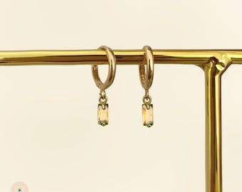 Puede incluir: Un par de pendientes de aro dorados con colgantes rectangulares de piedras preciosas iridiscentes. Los pendientes se exhiben en un soporte dorado para joyas. La marca "ATHENA" es visible en la parte inferior izquierda.