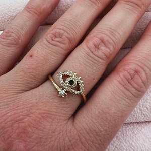 May include: Gold ring with a large eye design featuring black and white cubic zirconia stones. The eye is surrounded by smaller stones.