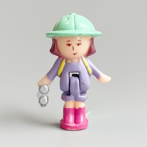 May include: A small plastic figurine of a girl wearing a green hat, purple jacket, pink boots, and a yellow backpack. The figurine is holding a pair of silver binoculars. The girl has red hair and is smiling.