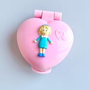 Puede incluir: Una caja de pl&aacute;stico rosa en forma de coraz&oacute;n con una peque&ntilde;a figura de mu&ntilde;eca de pl&aacute;stico en su interior. La mu&ntilde;eca lleva un vestido azul y tiene el pelo rubio. La caja tiene un peque&ntilde;o dise&ntilde;o de coraz&oacute;n en la parte delantera.