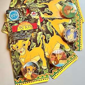 Könnte beinhalten: Eine Sammlung von Vintage Lion King POGs und Sammlerknöpfen. Die Verpackung zeigt Simba und andere Charaktere aus dem Zeichentrickfilm. Die Knöpfe haben verschiedene Designs, darunter Simba und andere Charaktere. Der Hintergrund hat ein Dschungelthema.