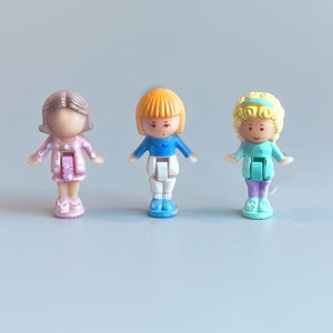 May include: Three plastic dolls with different colored hair and outfits. The doll on the left is wearing a pink and white dress, the middle doll is wearing a blue shirt and white pants, and the doll on the right is wearing a light blue dress.