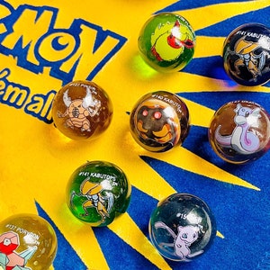 Puede incluir: Una alfombra azul y amarilla de Pokemon "catch 'em all" con nueve canicas de vidrio que representan diferentes personajes de Pokemon. Las canicas están dispuestas en un círculo alrededor del centro de la alfombra.