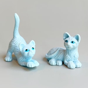 Könnte beinhalten: Zwei hellblaue Katzenfiguren mit blauen Augen. Eine Katze befindet sich in einer spielerischen Pose, mit gewölbtem Rücken und erhobenem Schwanz. Die andere Katze liegt. Beide Figuren haben detaillierte Fellstrukturen.