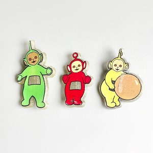 May include: Three Teletubbies figurines, one green, one red, and one yellow, standing on a white background. The yellow Teletubby is holding a round, orange object.