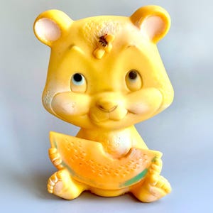 Op de afbeelding: Een geel rubberen speelgoeddier dat een plak watermeloen vasthoudt. Het speelgoed heeft een vriendelijk gezicht met grote ogen en een kleine neus.