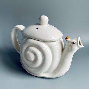 Puede incluir: Tetera de cerámica blanca con forma de caracol, con tapa y boquilla. La tetera tiene un diseño de espiral en la concha.
