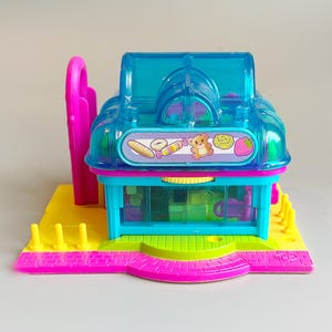 Puede incluir: Un colorido juego de panadería de juguete con un techo azul transparente, detalles en rosa y una base amarilla. La parte delantera presenta un mostrador con artículos en miniatura visibles. Un arco rosa está a la izquierda. El juego tiene una pegatina con un hámster, pan y una fresa.