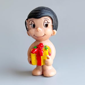 May include: A small figurine of a cartoon boy with black hair, holding a yellow gift box tied with a red ribbon. The figurine has a smiling expression and is standing on a gray surface.