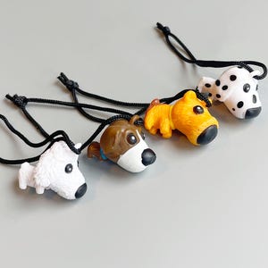 May include: Four small dog figurines in white, brown, yellow, and dalmatian patterns, each with a black cord loop. The dogs are lying down, with black noses and eyes. The background is a solid gray.