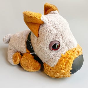 May include: A plush dog toy in a lying position. The toy has a light gray body with orange accents on the ears, paws, and muzzle. It has large, red-rimmed eyes and a black collar.