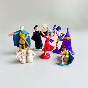 May include: A set of seven plastic figurines depicting characters from the Disney movie The Hunchback of Notre Dame. The characters include Quasimodo, Esmeralda, Frollo, Phoebus, Clopin, and two gargoyles. The figurines are painted in bright colors and are in good condition.