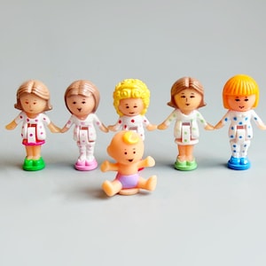 Può includere: Una collezione di sei piccole statuette di plastica. Cinque figure indossano abiti bianchi con pois colorati e scarpe abbinate. Una statuetta di un bambino è al centro, con un pannolino. Le figure sono disposte su una superficie grigia.