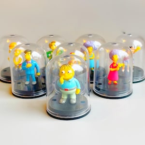 Puede incluir: Colección de figuras en miniatura de personajes de una popular serie de televisión animada, cada una encerrada en una cúpula de plástico transparente. Las figuras son de colores brillantes y se encuentran sobre bases negras. Las cúpulas están dispuestas sobre una superficie blanca.