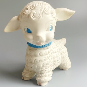 Peut inclure: Un agneau en caoutchouc blanc avec des yeux bleus et un ruban bleu autour du cou. L'agneau a un corps bouclé et texturé.