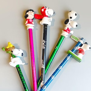 Op de afbeelding: Een verzameling kleurrijke potloden met Snoopy en andere Peanuts-personages. De potloden hebben verschillende ontwerpen, waaronder Snoopy met een voetbal en Lucy in een rode jurk. De potloden zijn van Applause, Woodland Hills, CA 91365.