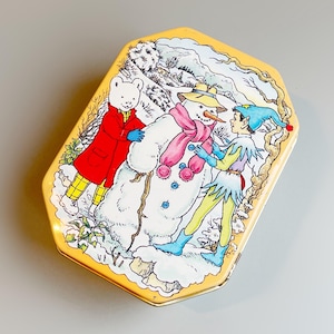 Puede incluir: Una caja de lata vintage con borde dorado, que representa una escena invernal con un muñeco de nieve, un oso con abrigo rojo y un personaje con sombrero azul y atuendo verde. La caja es rectangular con bordes redondeados y una ilustración detallada.