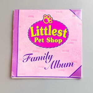 May include: A pink "Littlest Pet Shop Family Album" with a purple spine and accents. The cover features a pink oval with the words "Littlest Pet Shop" in yellow and a paw print. The words "Family Album" are written in cursive.