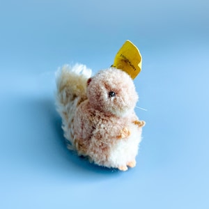 May include: A small, plush squirrel toy with a white tail and a pink body. The toy has a yellow tag that says "Steiff" on it.