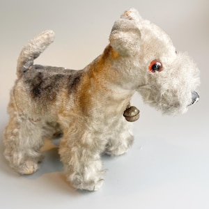 Può includere: Un giocattolo vintage a forma di cane di peluche con un collare a campana. Il cane ha un corpo soffice e chiaro con segni marroni e grigi. Ha occhi rossi e un piccolo naso nero. Il cane è in piedi su tutte e quattro le zampe.