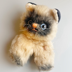 Op de afbeelding: Een vintage pluche kattenspeelgoed met een crèmekleurig lichaam en een donkerbruin gezicht en oren. De kat heeft één blauw oog en één zwart oog, met een oranje neus en snorharen. Het speelgoed is gemaakt van zacht, pluizig materiaal.